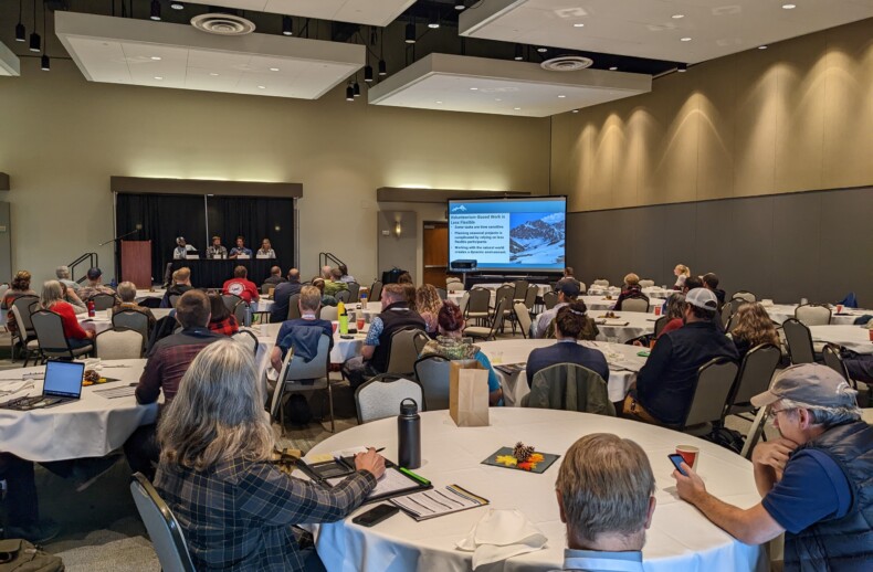 A crowd of conference attendees sit around large circular tables in a conference room. A panel of four presenters are seated along a raised stage at the back wall with a slideshow presentation projected onto a nearby screen.