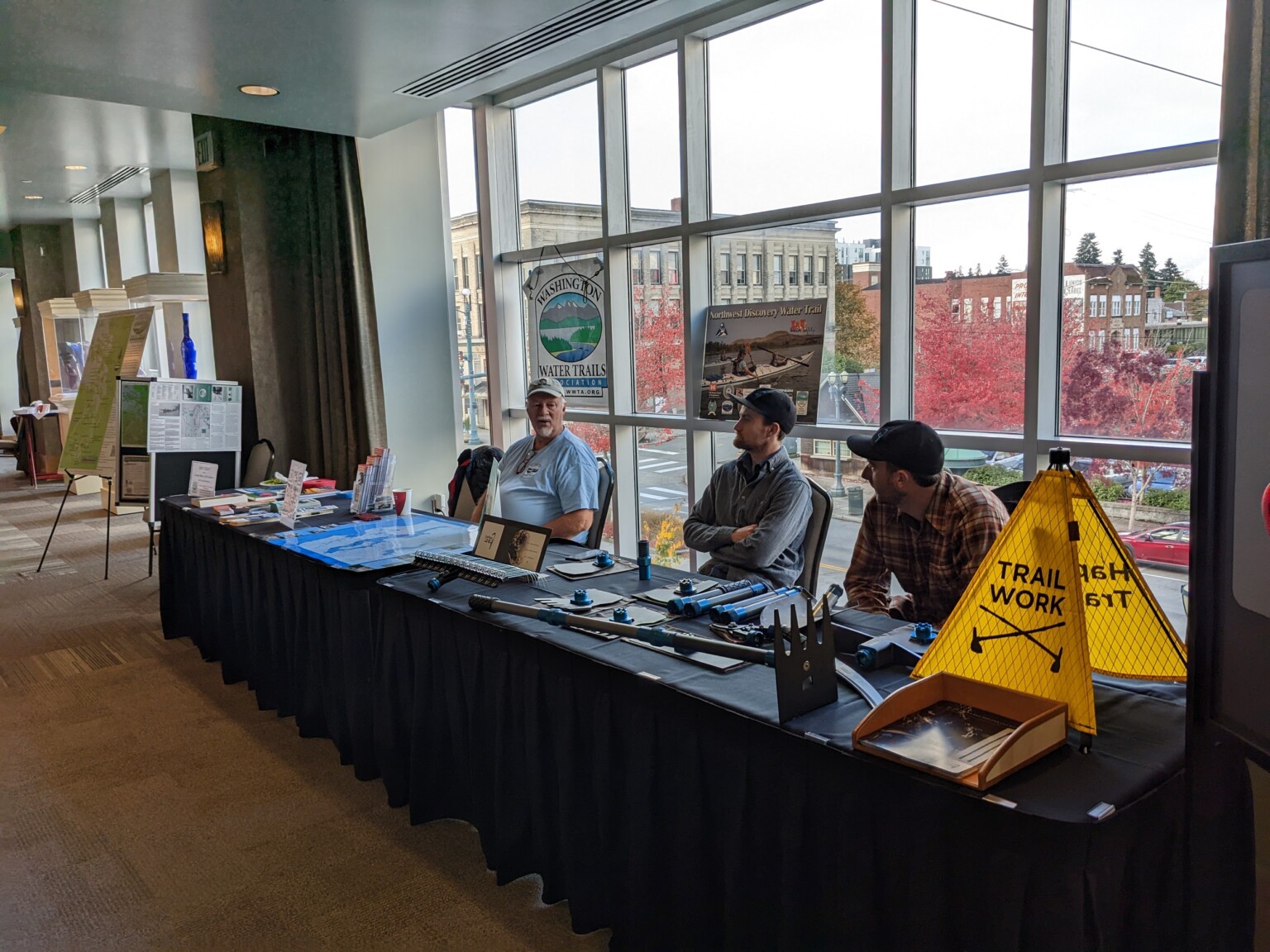 A sponsor table set up in a conference hallway with assorted paper materials spread out on top of it and several people sitting behind it.