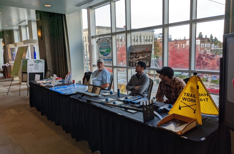 A sponsor table set up in a conference hallway with assorted paper materials spread out on top of it and several people sitting behind it.